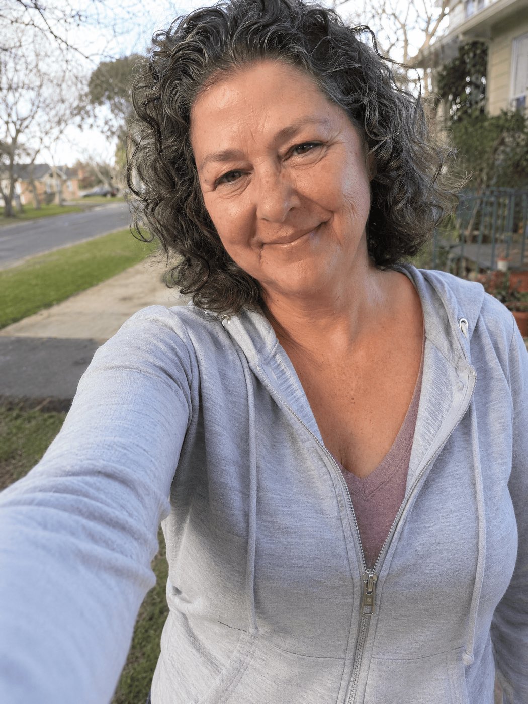 A natural, slightly imperfect selfie-style outdoor portrait of a happy, healthy woman ~54 with a slim face, real skin and a warm genuine smile, wearing a casual zip-up jacket, with a suburban street slightly blurred behind her. Looks like a real photo a real woman would post.